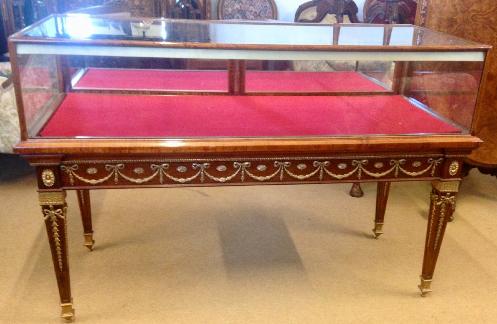 An antique glazed mahogany bijouterie table with gilt brass swags, square tapered legs, glass panels, and a red velvet interior display lining.