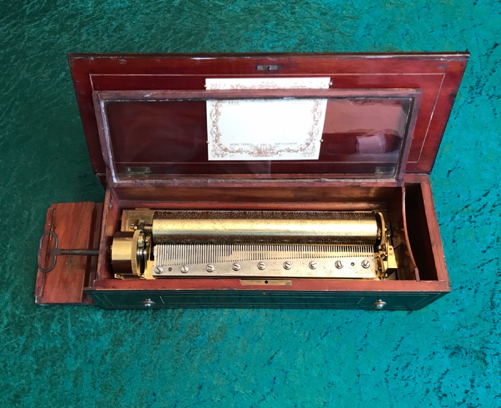 A rectangular dark wood music box with floral marquetry inlay on the lid and a metal winding key resting on top. View V