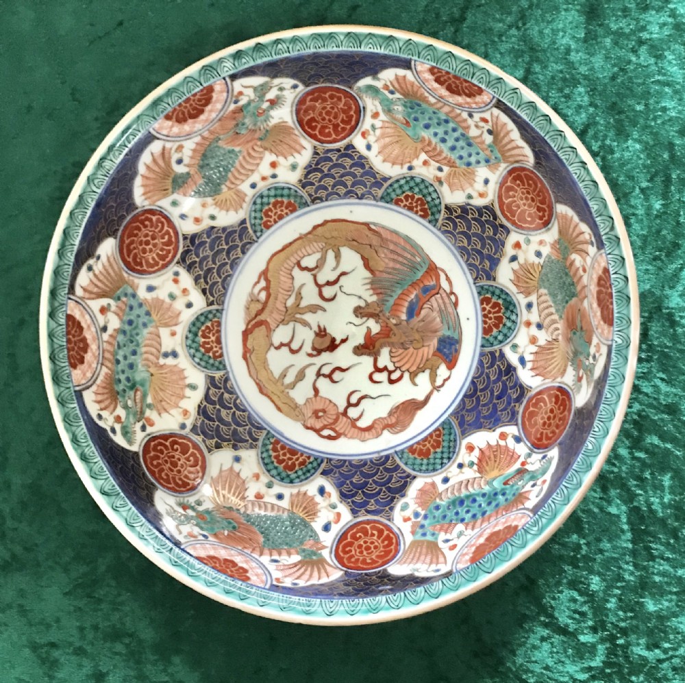 A circular antique Japanese Imari porcelain bowl decorated with a central dragon medallion surrounded by hand-painted fish, wave patterns, and floral motifs in blue, terracotta red, and turquoise.