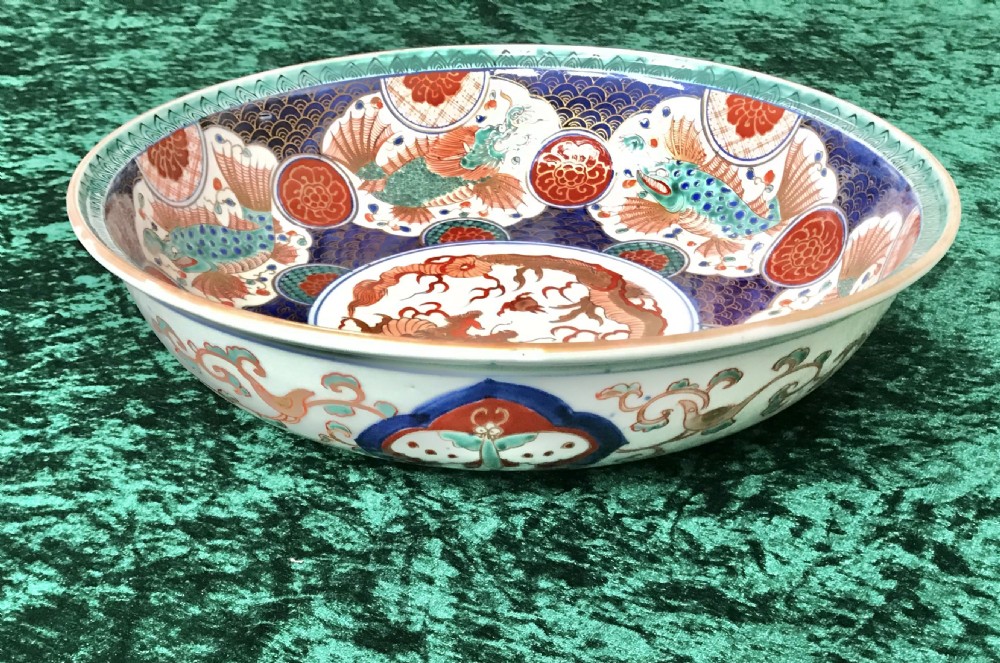 A circular antique Japanese Imari porcelain bowl decorated with a central dragon medallion surrounded by hand-painted fish, wave patterns, and floral motifs in blue, terracotta red, and turquoise. View II
