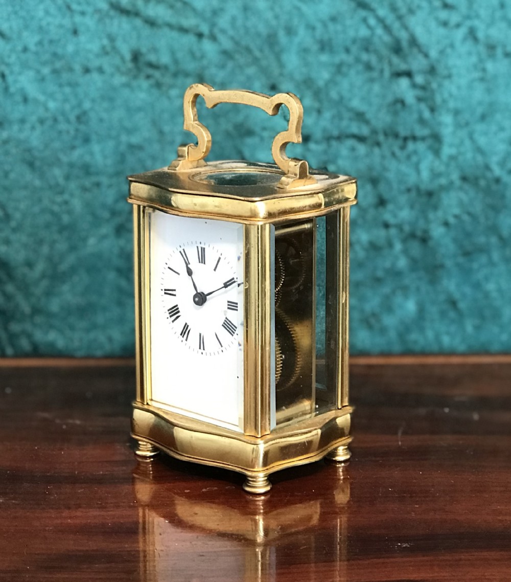 An antique brass serpentine-cased carriage clock featuring glass side panels, a white enamel dial with black Roman numerals, and a top carrying handle.