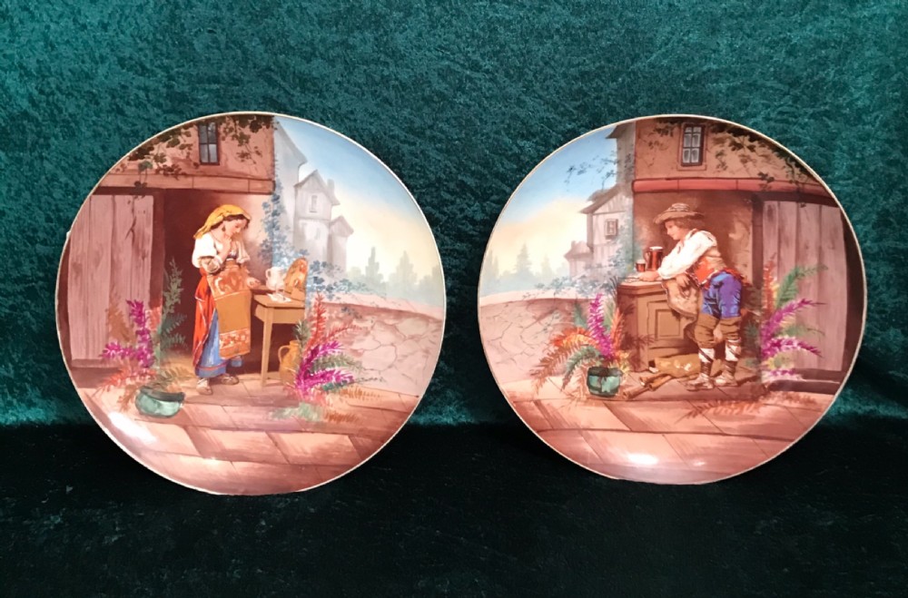 A pair of circular ceramic wall plaques depicting hand-painted European rustic genre scenes with figures in traditional dress against village backdrops.