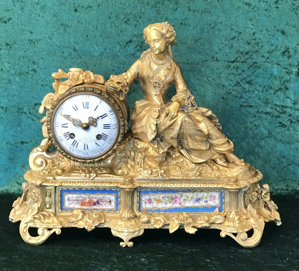 A 19th-century French gilt bronze mantel clock featuring a sculpted reclining female figure beside a round dial mounted on an ornate base with porcelain plaques.
