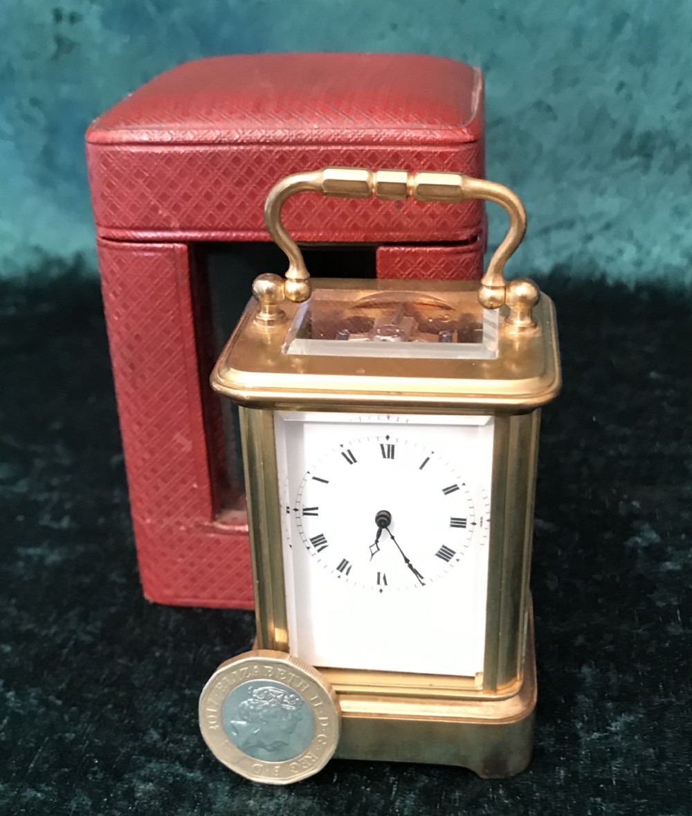 A miniature brass carriage clock with a white enamel dial and Roman numerals, standing beside its fitted red leather travel case.