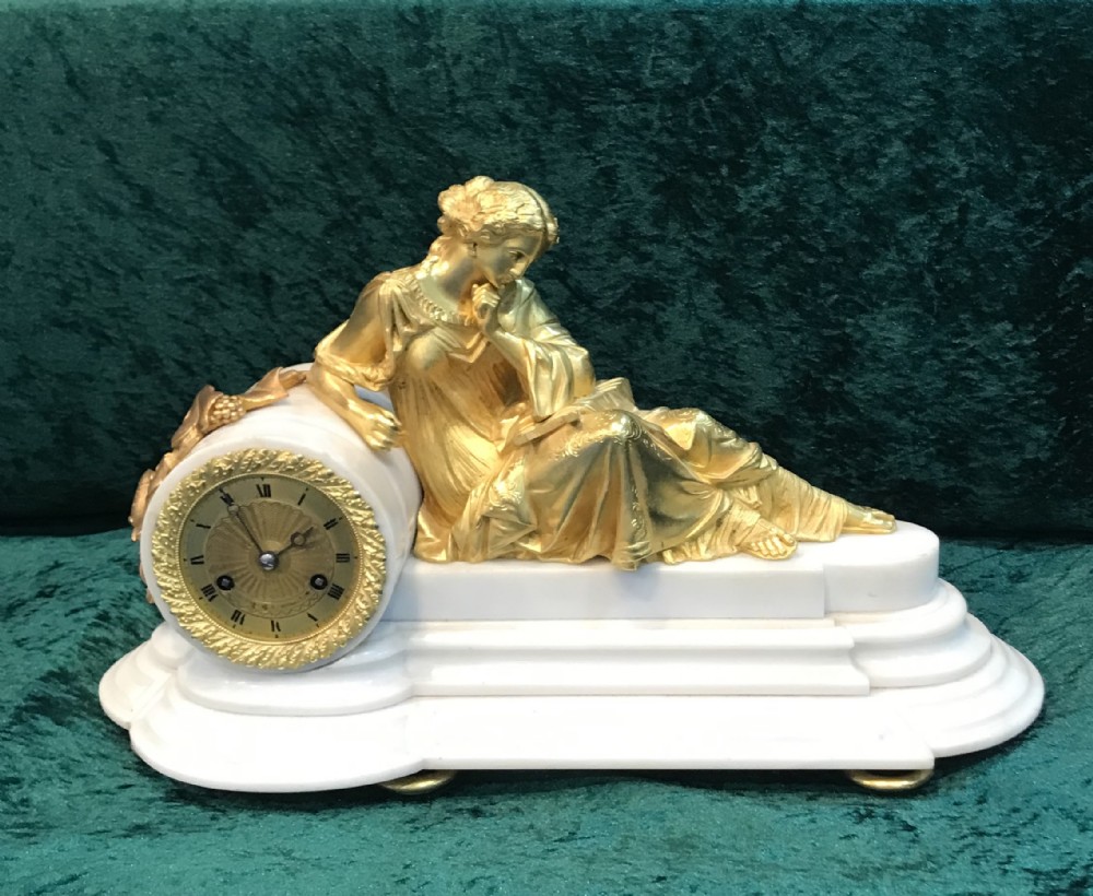 A Victorian gilt bronze and white marble mantel clock featuring a reclining classical female figure reading a book atop a stepped base.