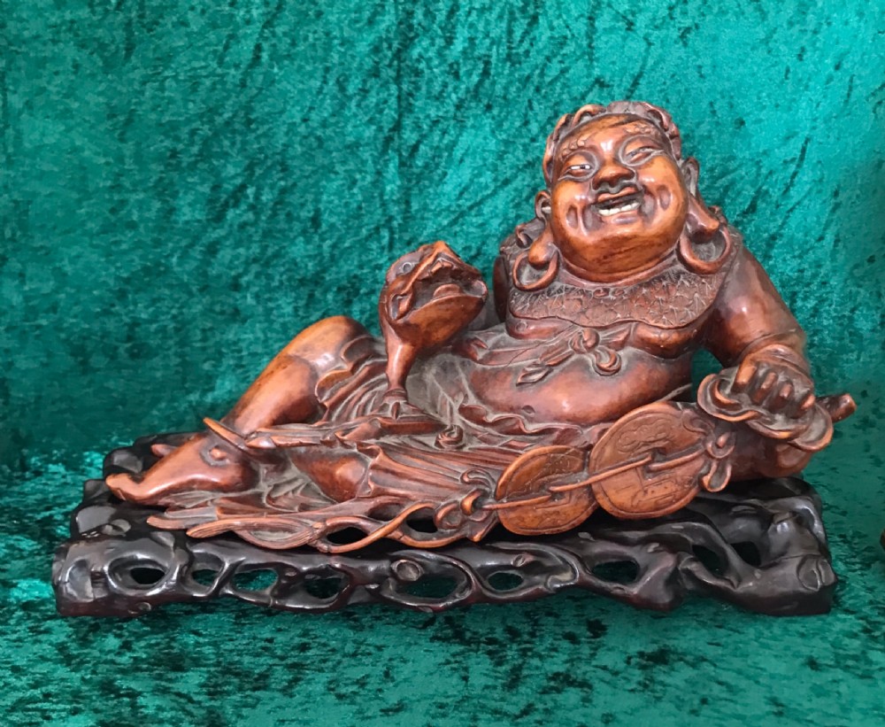 A carved warm brown wooden figure of a reclining laughing Buddha seated on a pierced dark hardwood stand holding a string of coins alongside a frog.