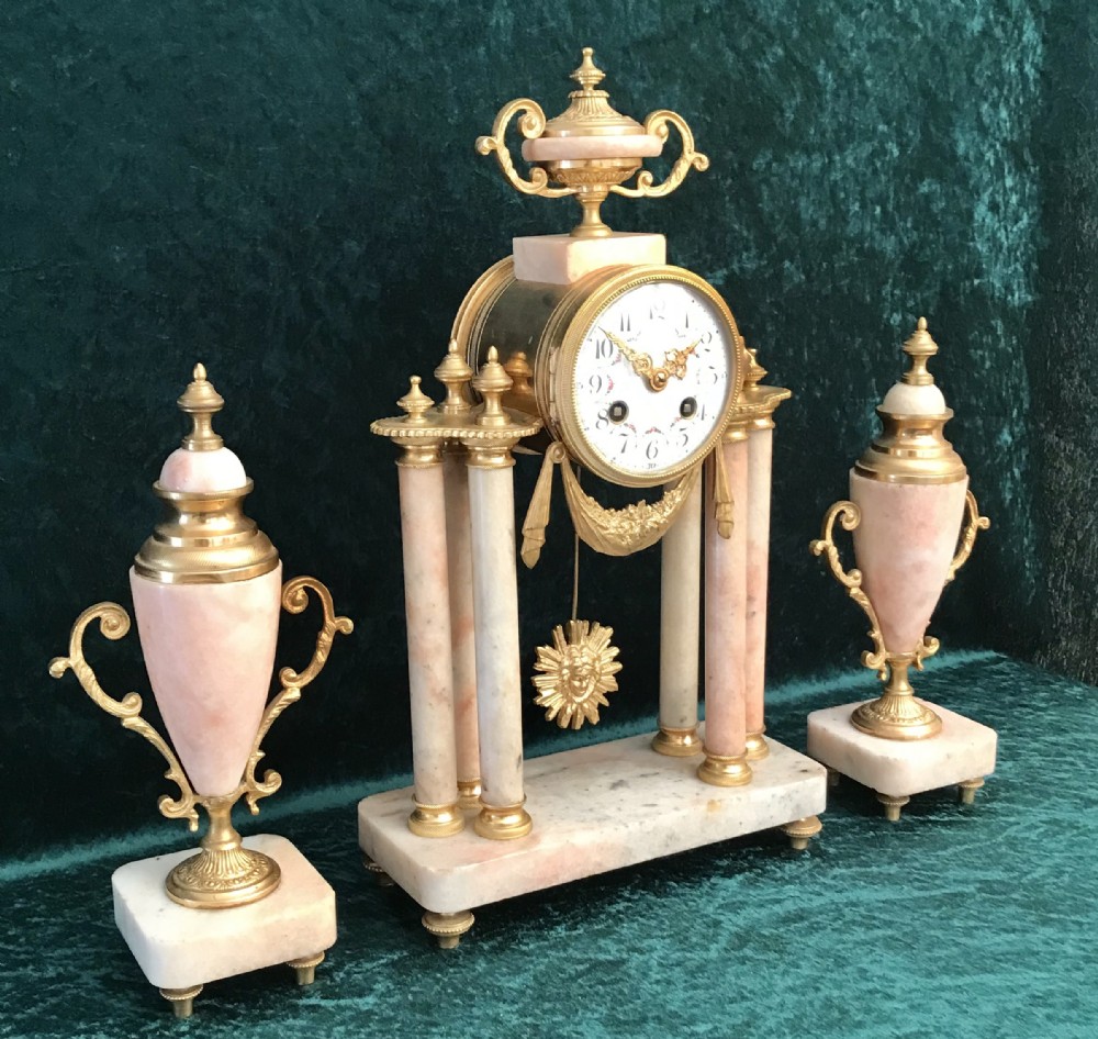 A nineteenth-century pink marble portico clock supported by four columns and gilded mounts, flanked by two matching ornamental urn garnitures.