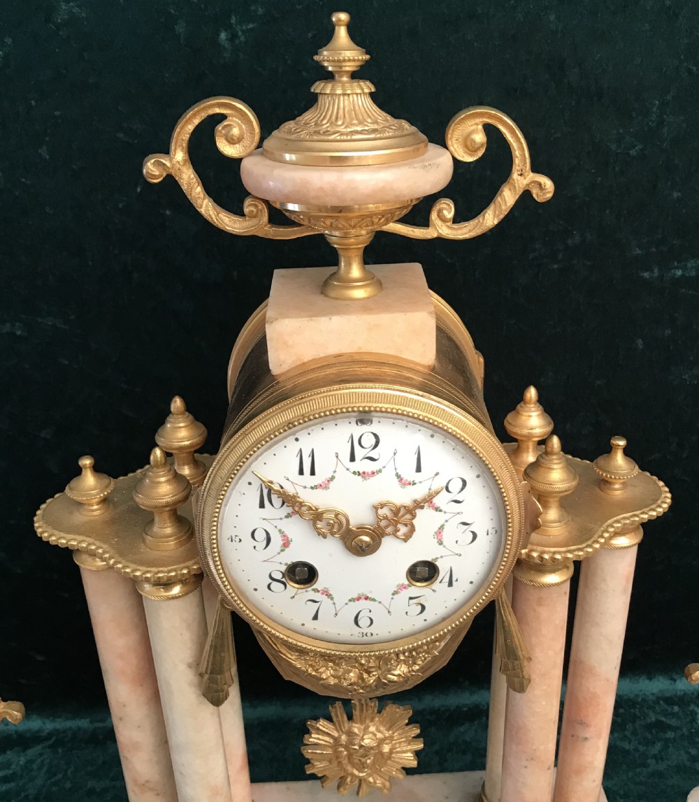 A nineteenth-century pink marble portico clock supported by four columns and gilded mounts, flanked by two matching ornamental urn garnitures. View III