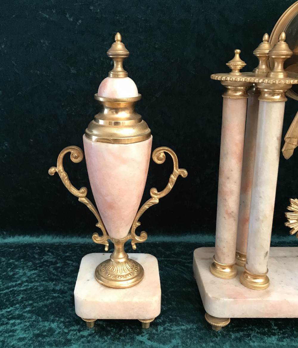 A nineteenth-century pink marble portico clock supported by four columns and gilded mounts, flanked by two matching ornamental urn garnitures. View VI