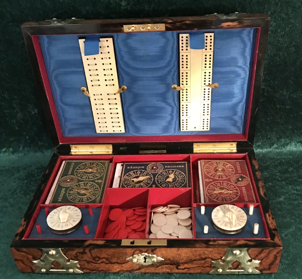 An open Victorian burr walnut games box with brass corner mounts, revealing a silk-lined lid with scoring boards and red fitted compartments filled with card registers and gaming counters.