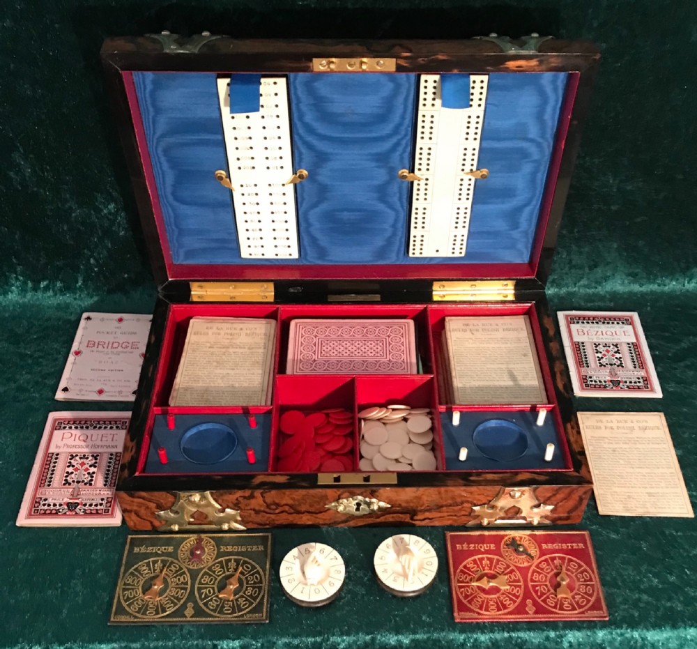 An open Victorian burr walnut games box with brass corner mounts, revealing a silk-lined lid with scoring boards and red fitted compartments filled with card registers and gaming counters. View VI