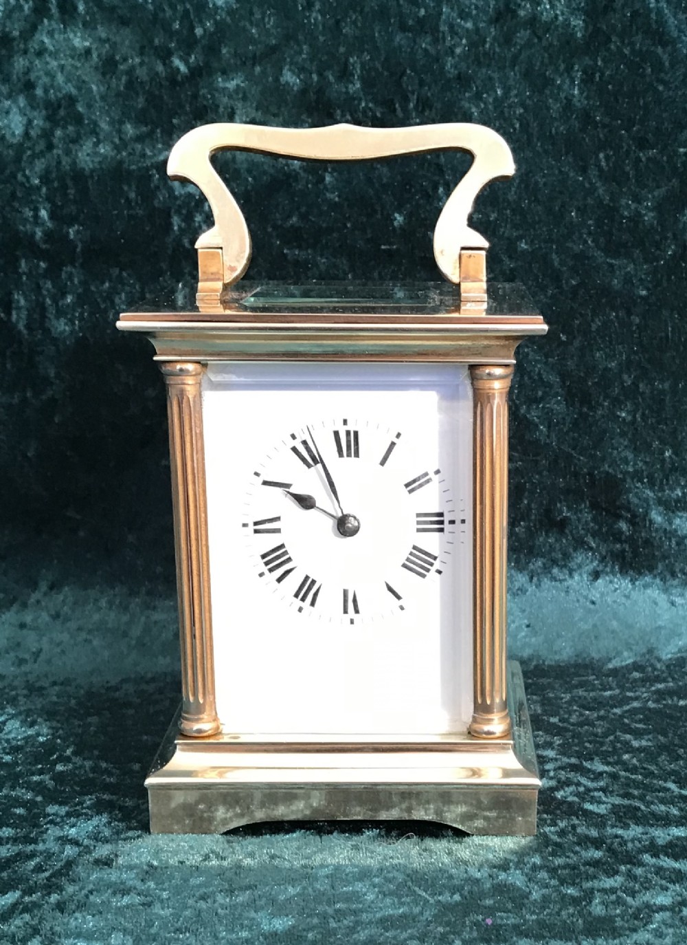 An antique brass carriage clock featuring a top carrying handle, reeded corner columns, and a white enamel dial with black Roman numerals.