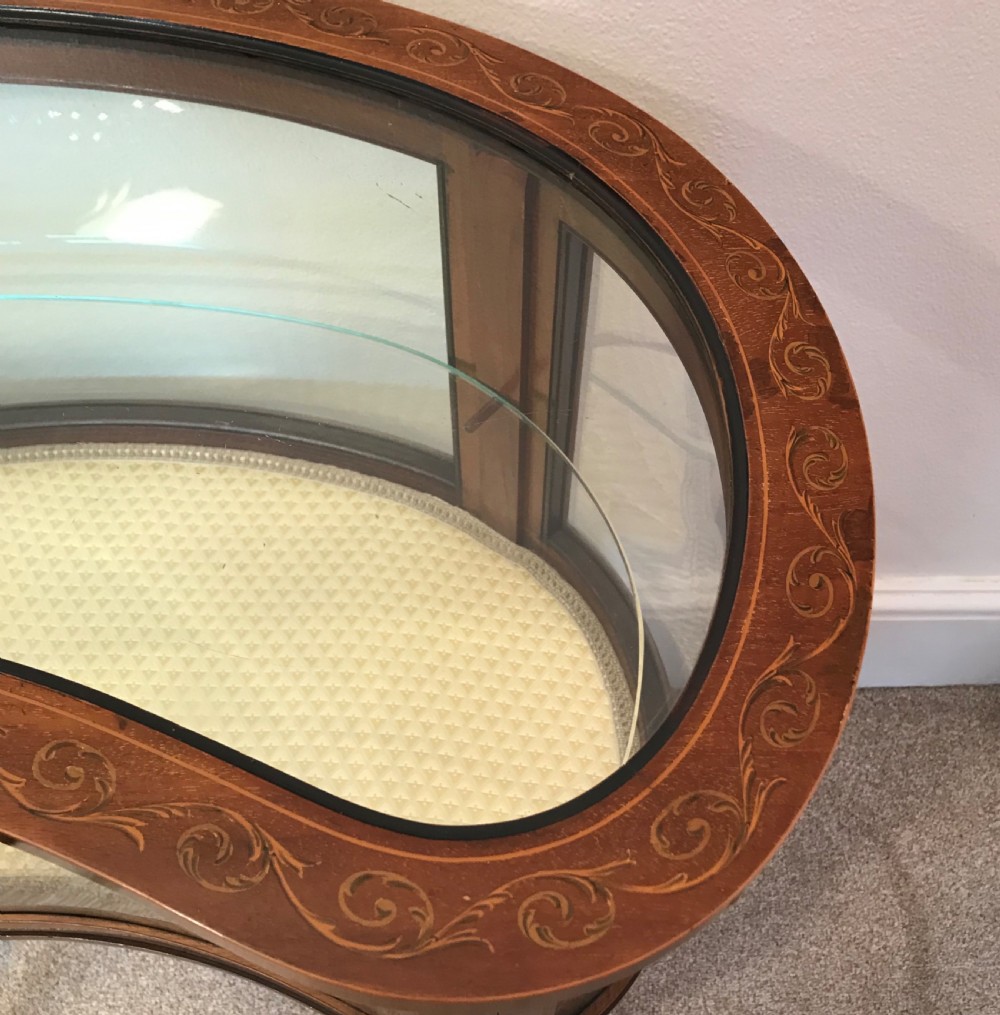 An Edwardian kidney-shaped mahogany bijouterie table with glazed top and sides, floral inlay, slender curved legs, and an interior glass shelf. View V