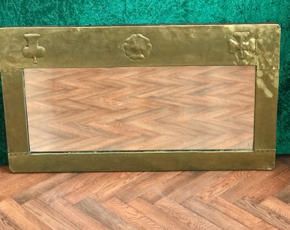 A large rectangular Arts and Crafts wall mirror featuring a hand-hammered brass frame decorated with repousse thistle, rose, and shamrock motifs.