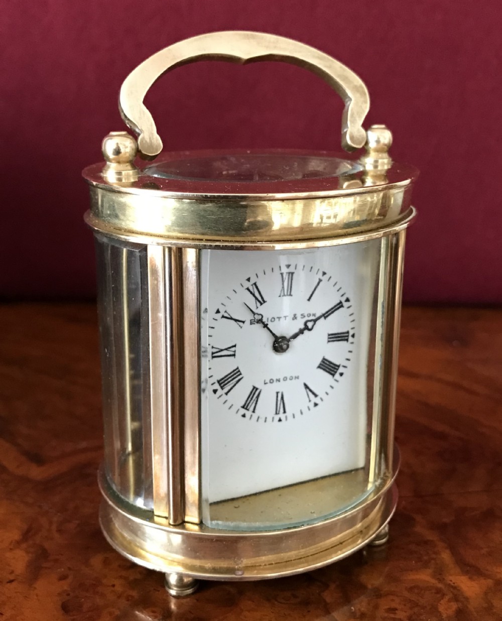 A miniature oval brass carriage clock featuring glass panels, a top handle, and a white dial with Roman numerals signed Elliott and Son London.