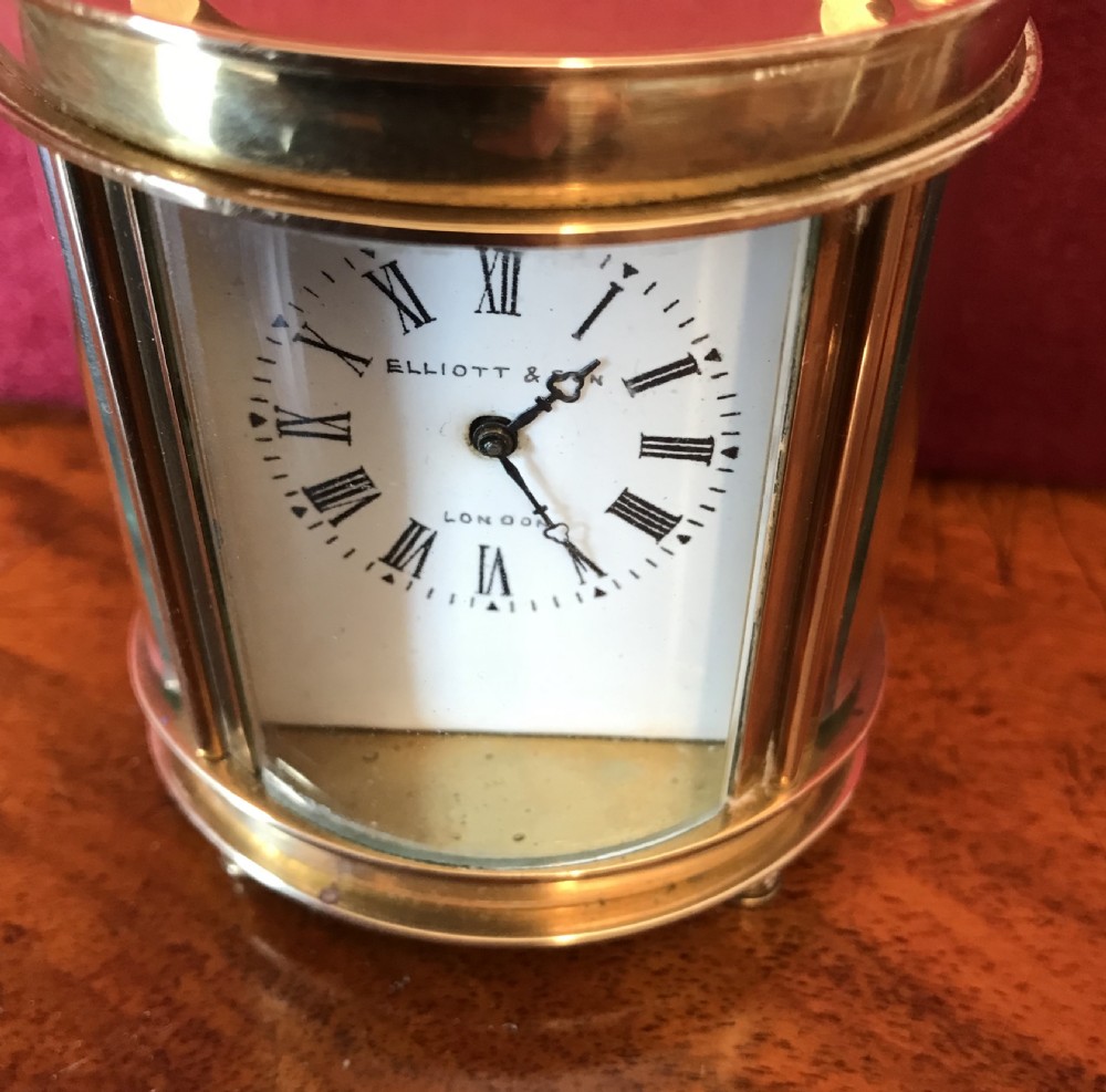 A miniature oval brass carriage clock featuring glass panels, a top handle, and a white dial with Roman numerals signed Elliott and Son London. View III