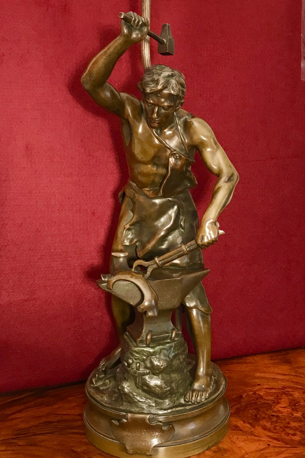 A patinated bronze figural table lamp depicting a muscular blacksmith standing at an anvil holding a hammer and tongs, raised on a moulded circular base.