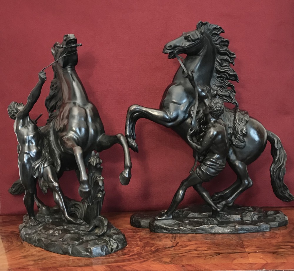 A pair of patinated dark bronze sculptures depicting rearing horses held by grooms on naturalistic bases.
