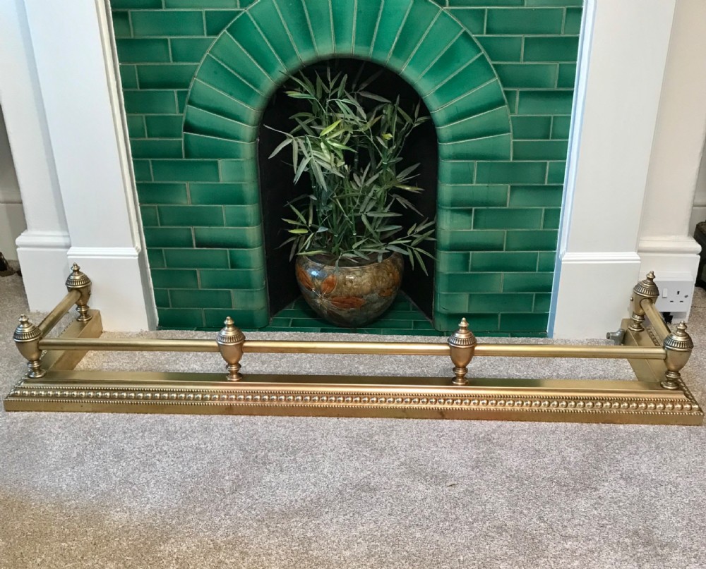 A heavy gauge Victorian brass fireplace fender featuring urn finials, a tubular top rail, and a beaded moulded base.