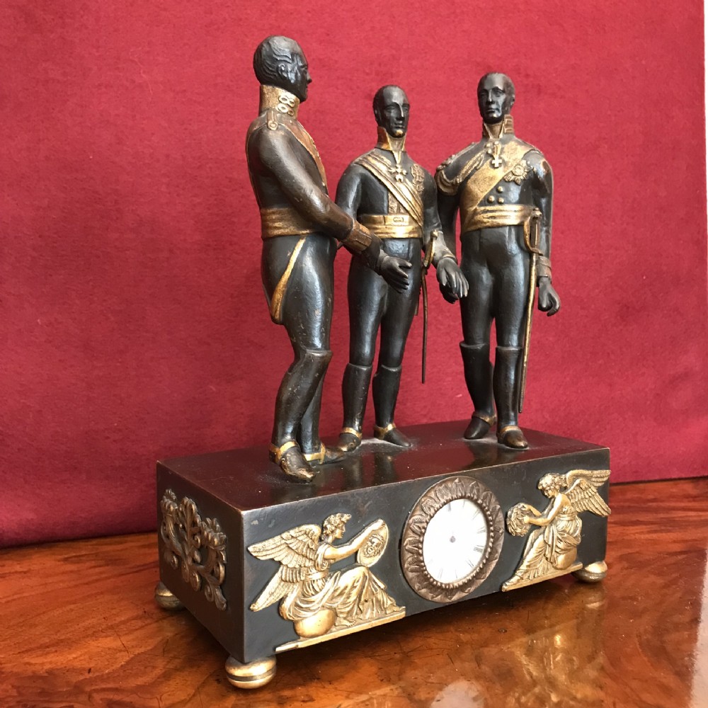 A dark patinated bronze clock featuring three standing military figures on a rectangular plinth with gilt-bronze relief mounts and a circular dial.