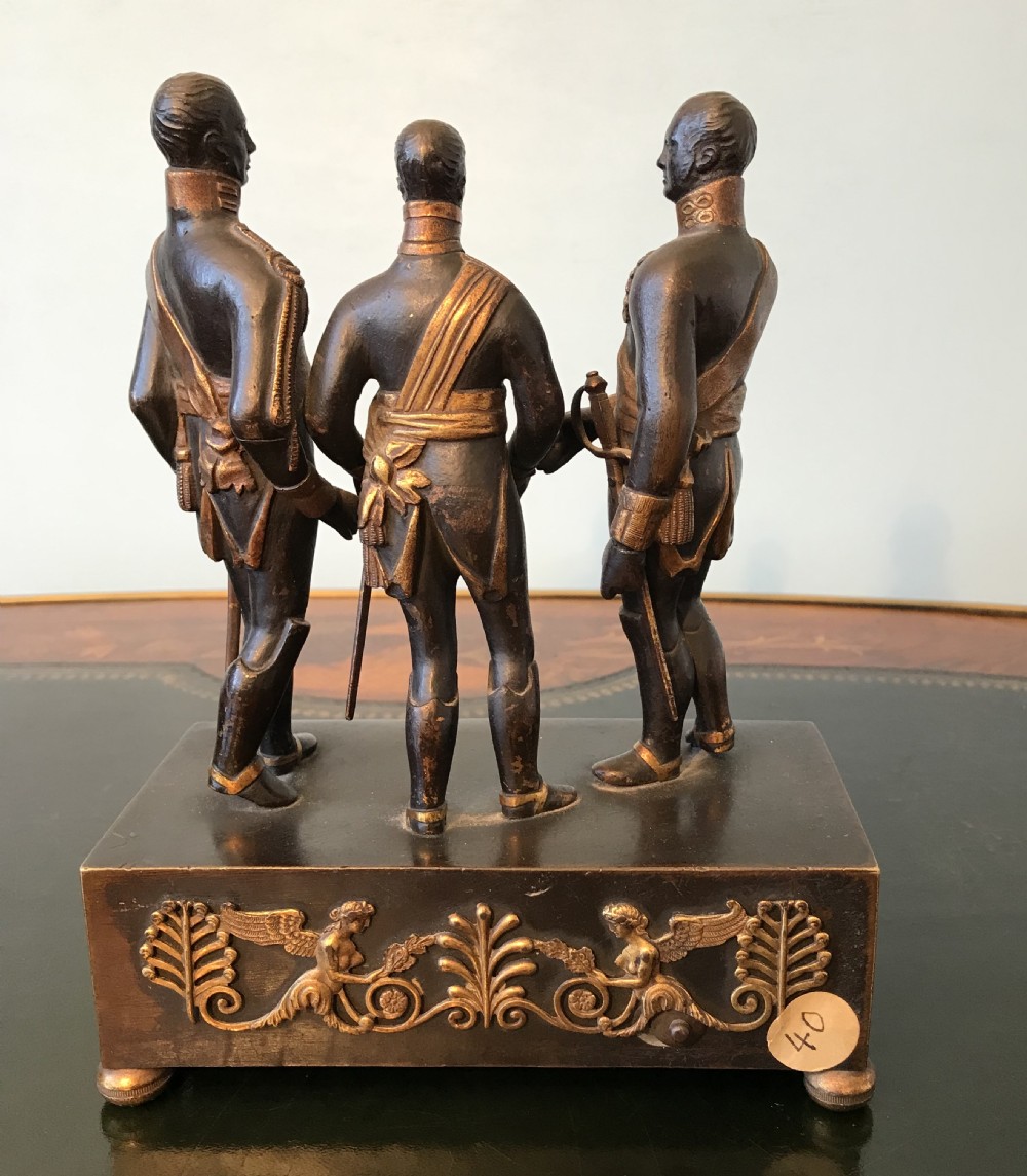 A dark patinated bronze clock featuring three standing military figures on a rectangular plinth with gilt-bronze relief mounts and a circular dial. View V