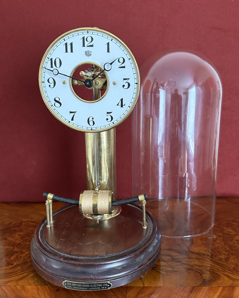 An antique Bulle electric clock featuring an exposed brass mechanism, white dial with Arabic numerals, mahogany circular base, and removable glass dome.