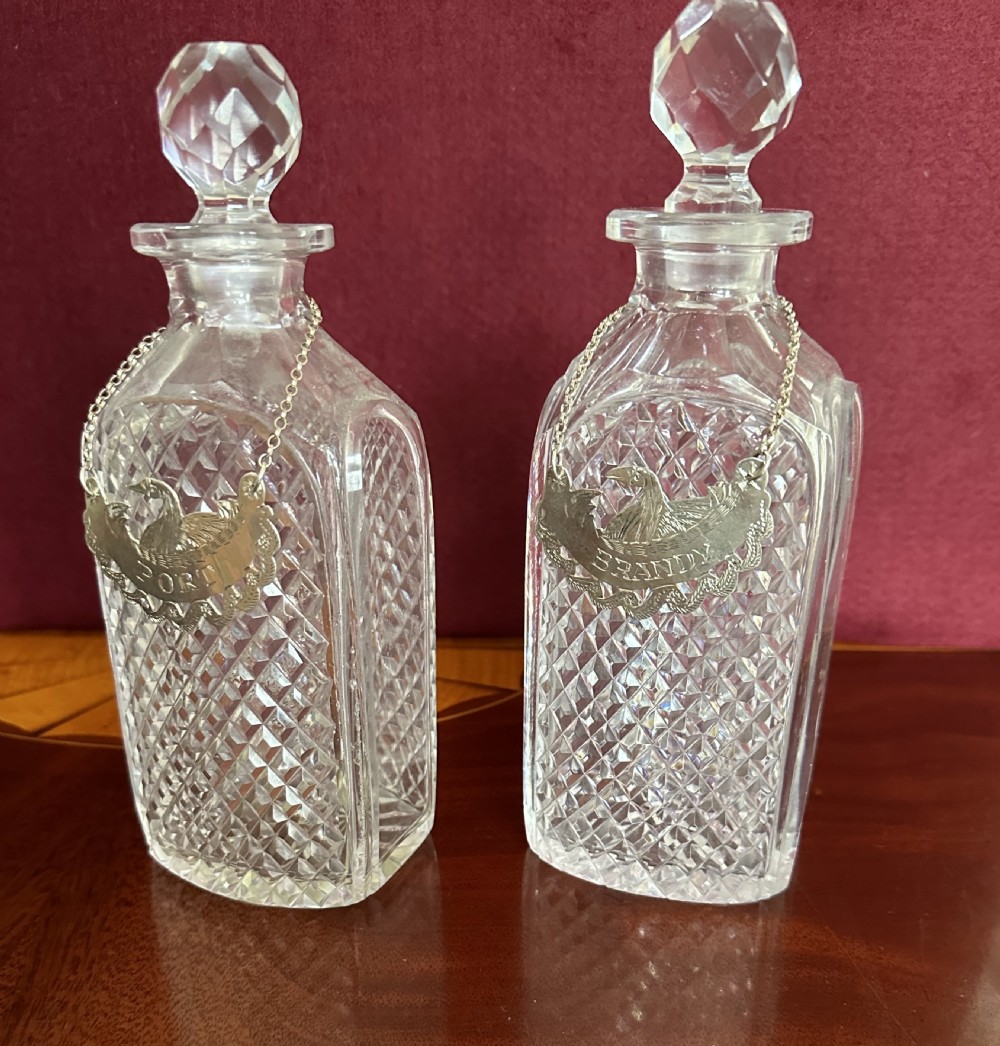 A pair of clear Edwardian cut glass decanters featuring diamond facets, faceted stoppers, and silver decanter labels on chains. View V