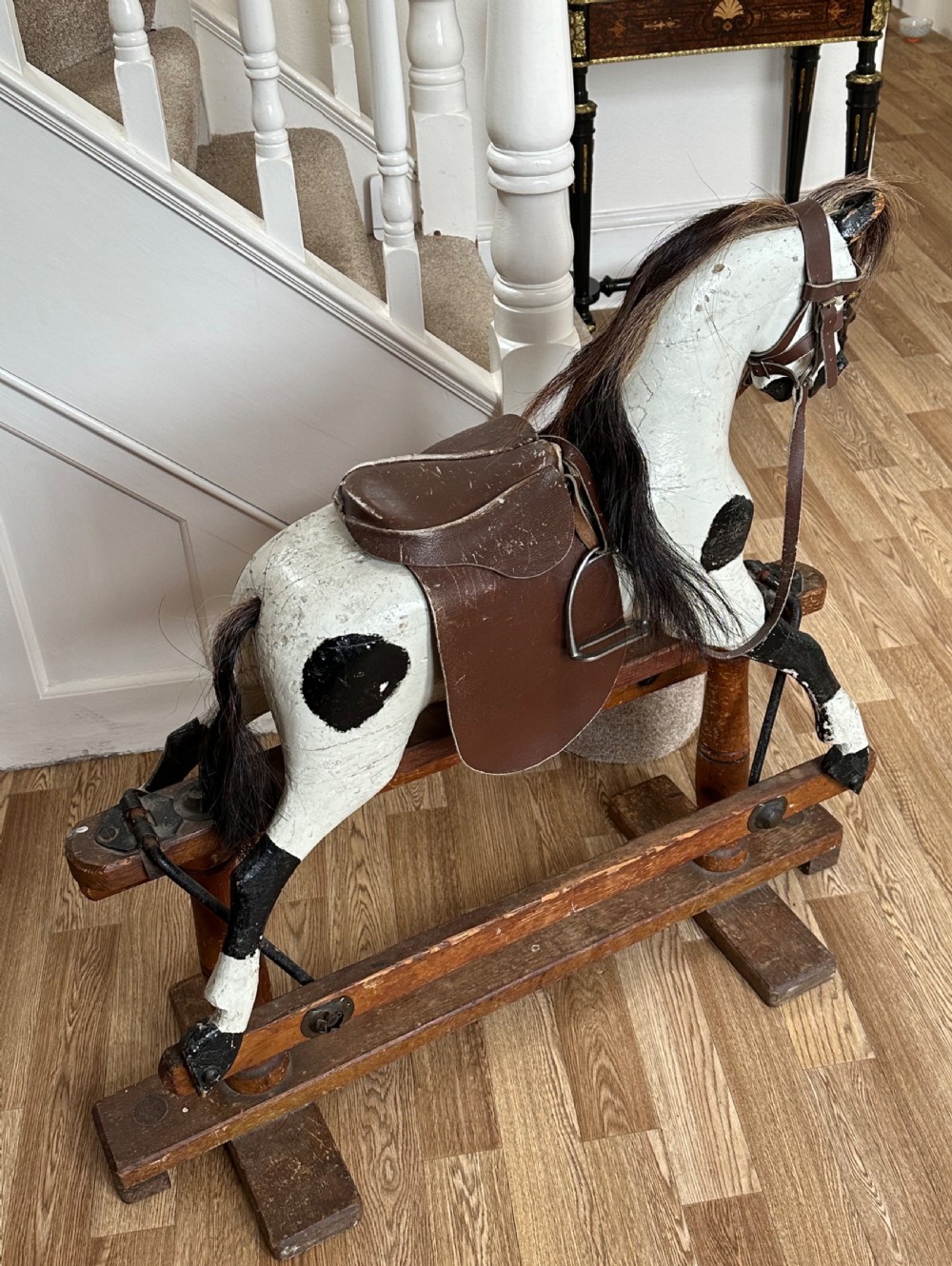 An antique carved wooden rocking horse with a white painted body featuring black dapple spots, a brown leather saddle, and a horse hair mane mounted on a wooden bow rocker stand. View III