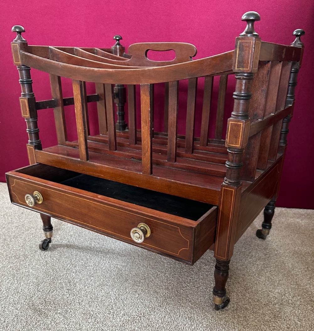 An antique mahogany music canterbury featuring slatted divisions, boxwood inlay, a single lower drawer with brass knobs, and turned legs on castors.