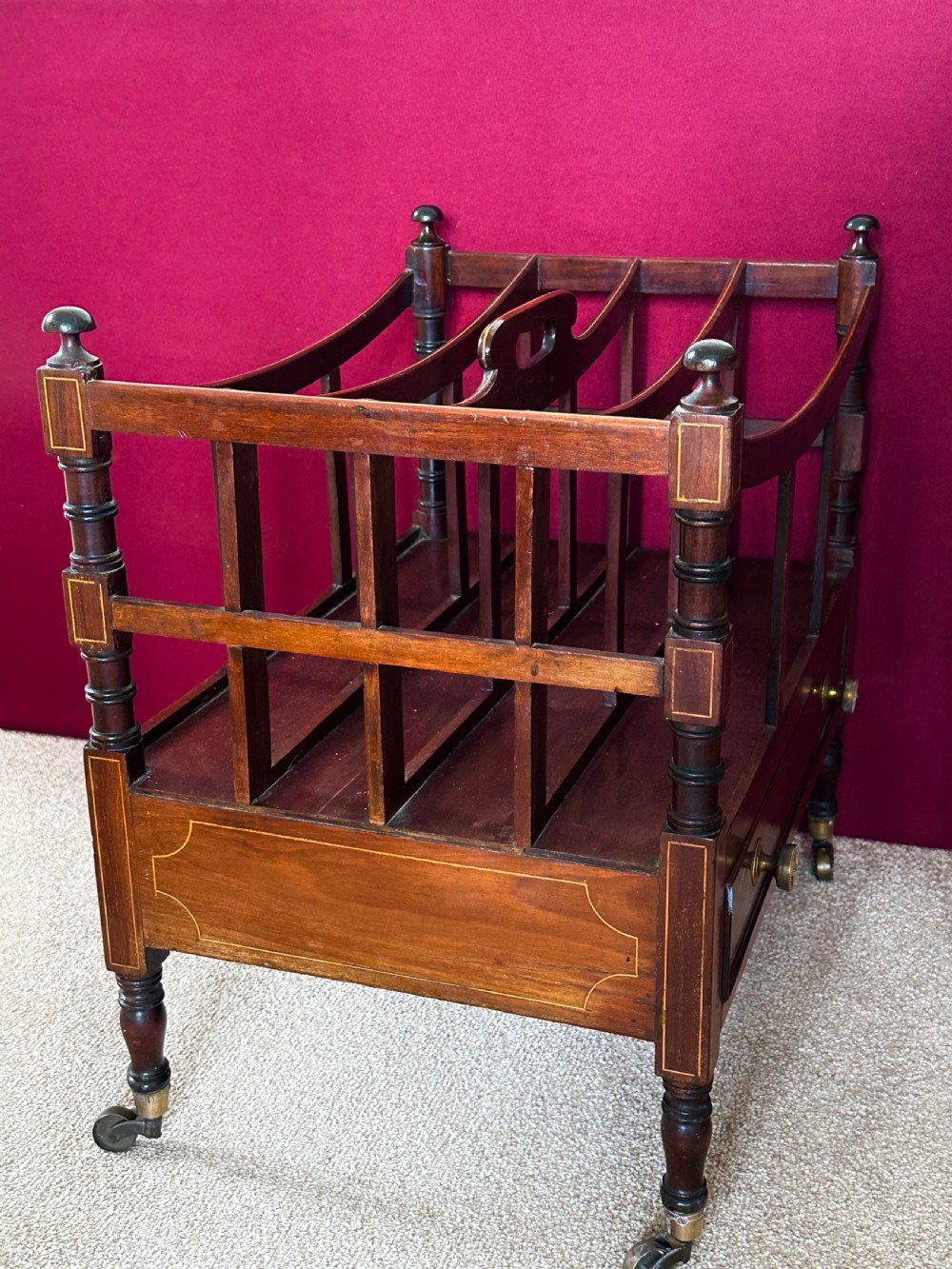 An antique mahogany music canterbury featuring slatted divisions, boxwood inlay, a single lower drawer with brass knobs, and turned legs on castors. View VI