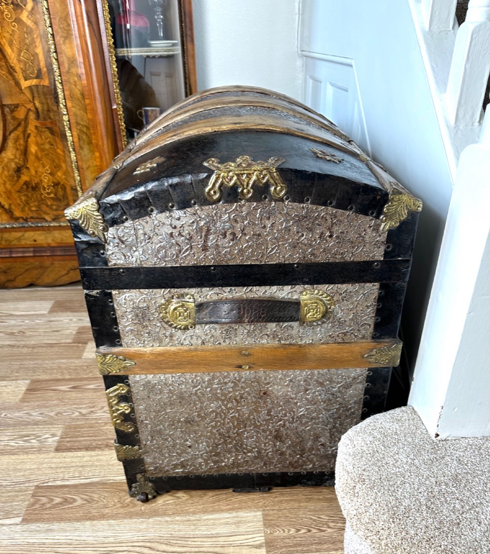 A Victorian dome top travelling trunk featuring embossed patterned metal casing, dark timber reinforcing slats, and brass corner mounts and central lock. View X