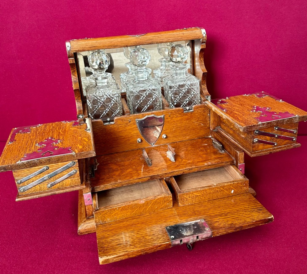 An opened Edwardian oak tantalus compendium box featuring three cut-glass decanters, a mirrored back, and hinged cantilever side compartments with silver-plated fittings. View III