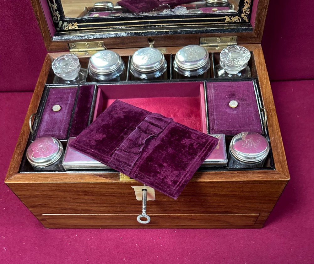 An open Victorian walnut travelling dressing case featuring an interior lid mirror, purple velvet compartments, glass jars with silver caps, and an opened lower drawer. View II
