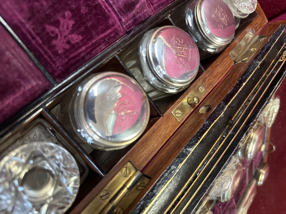 An open Victorian walnut travelling dressing case featuring an interior lid mirror, purple velvet compartments, glass jars with silver caps, and an opened lower drawer. View V
