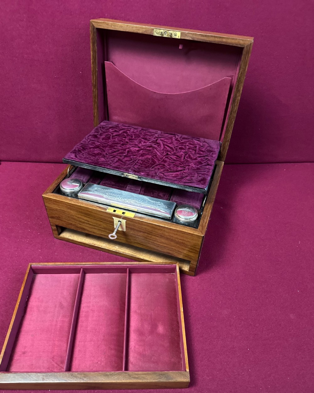 An open Victorian walnut travelling dressing case featuring an interior lid mirror, purple velvet compartments, glass jars with silver caps, and an opened lower drawer. View VI