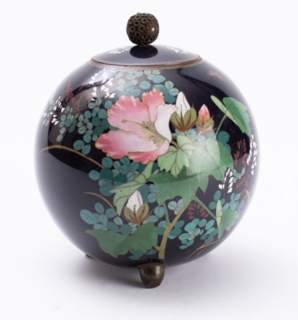 A spherical dark navy blue Japanese cloisonne enamel jar decorated with pink floral motifs, green foliage, three feet, and a lidded top with a filigree finial.
