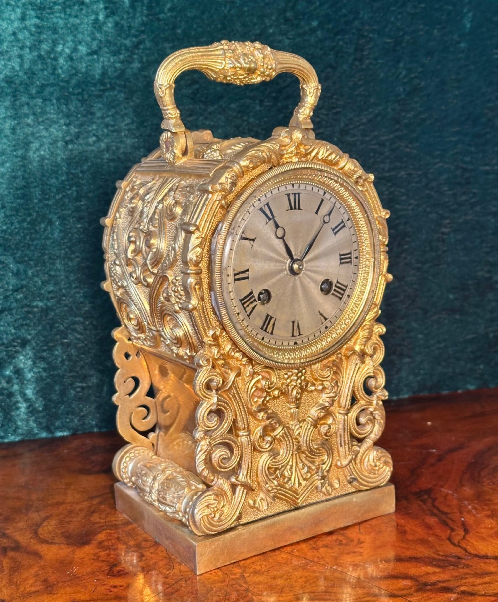An ornate gilt brass carriage clock featuring detailed Rococo scrollwork, a circular metal dial with Roman numerals, and an upper carrying handle.