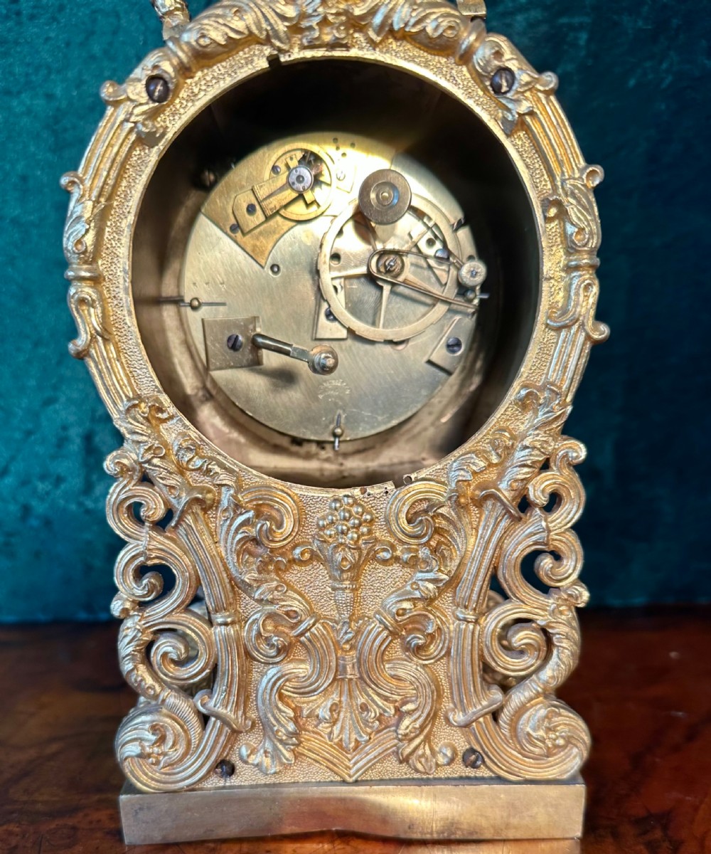 An ornate gilt brass carriage clock featuring detailed Rococo scrollwork, a circular metal dial with Roman numerals, and an upper carrying handle. View III