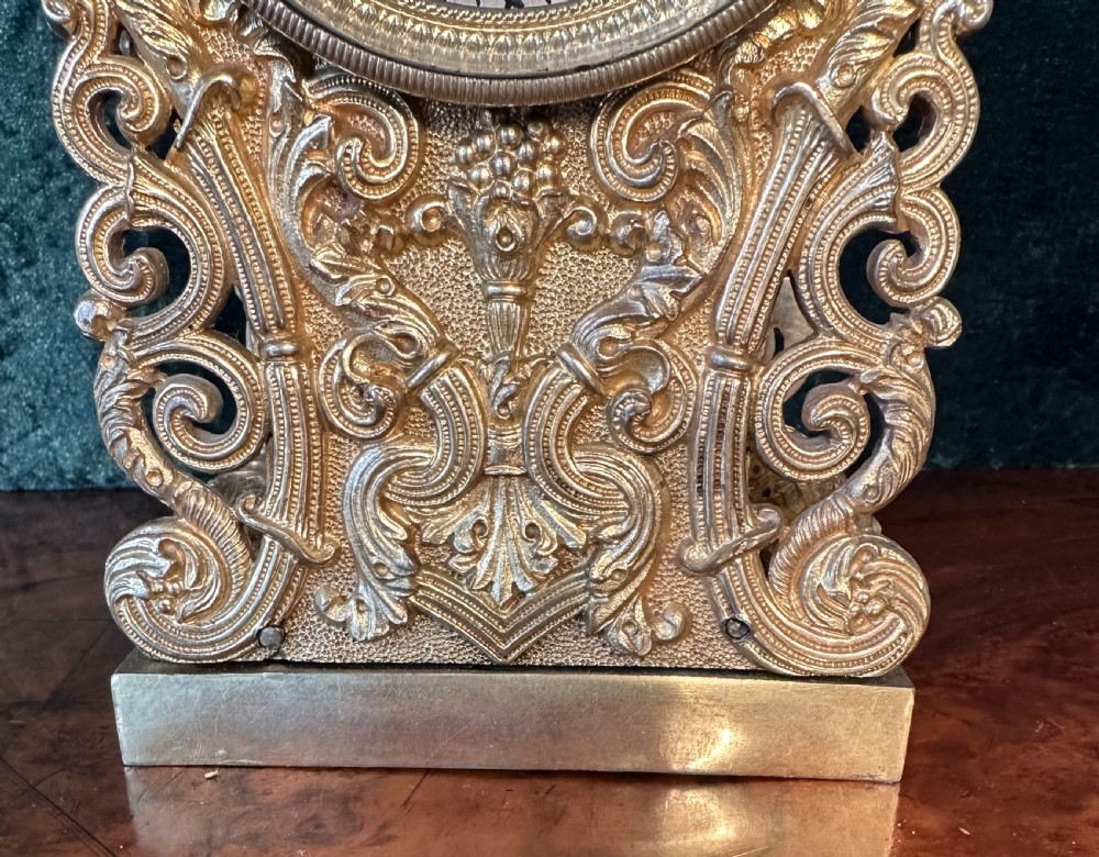 An ornate gilt brass carriage clock featuring detailed Rococo scrollwork, a circular metal dial with Roman numerals, and an upper carrying handle. View VIII