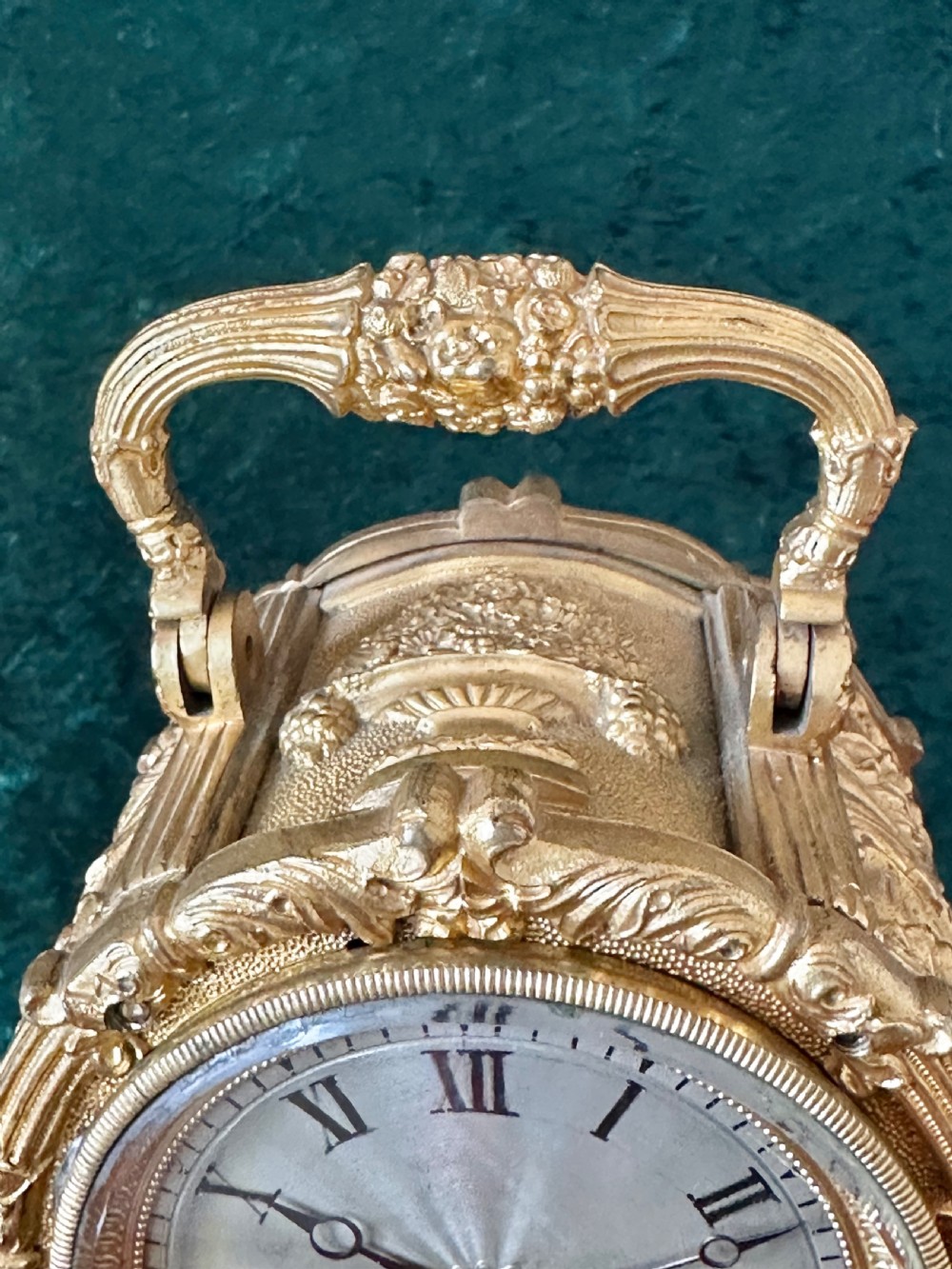 An ornate gilt brass carriage clock featuring detailed Rococo scrollwork, a circular metal dial with Roman numerals, and an upper carrying handle. View X