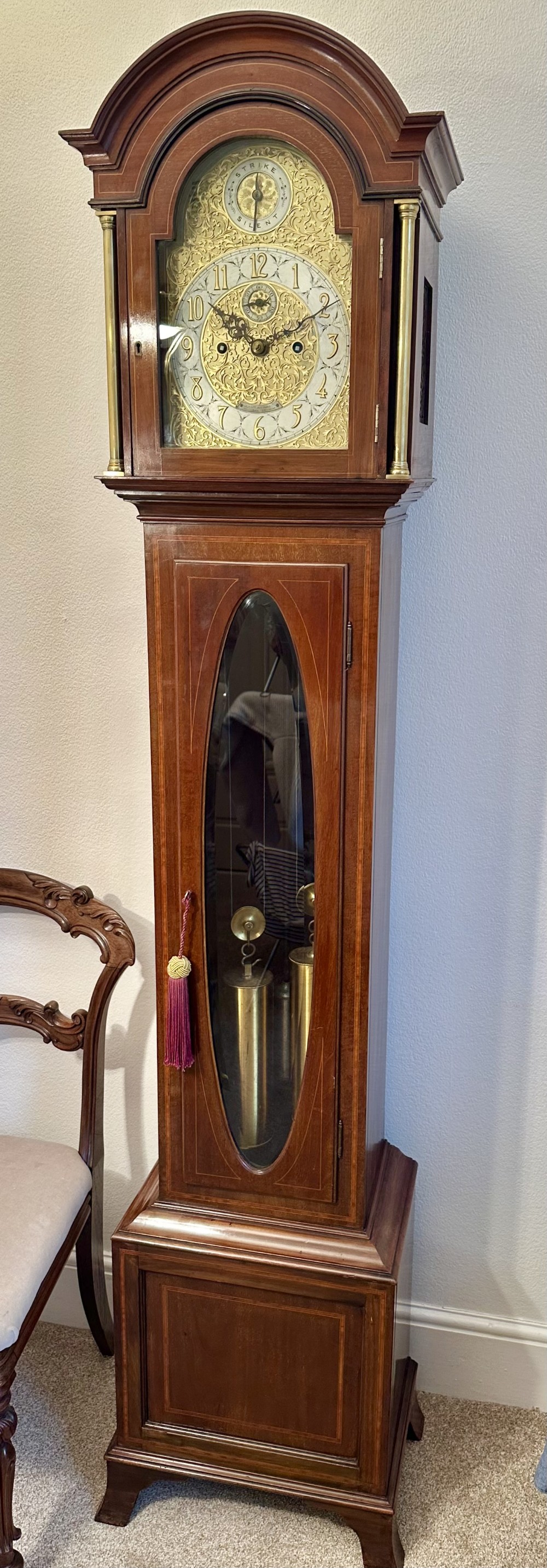 An Edwardian mahogany longcase clock featuring an arched hood, ornamental brass dial, and an oval glass aperture displaying the brass weights.