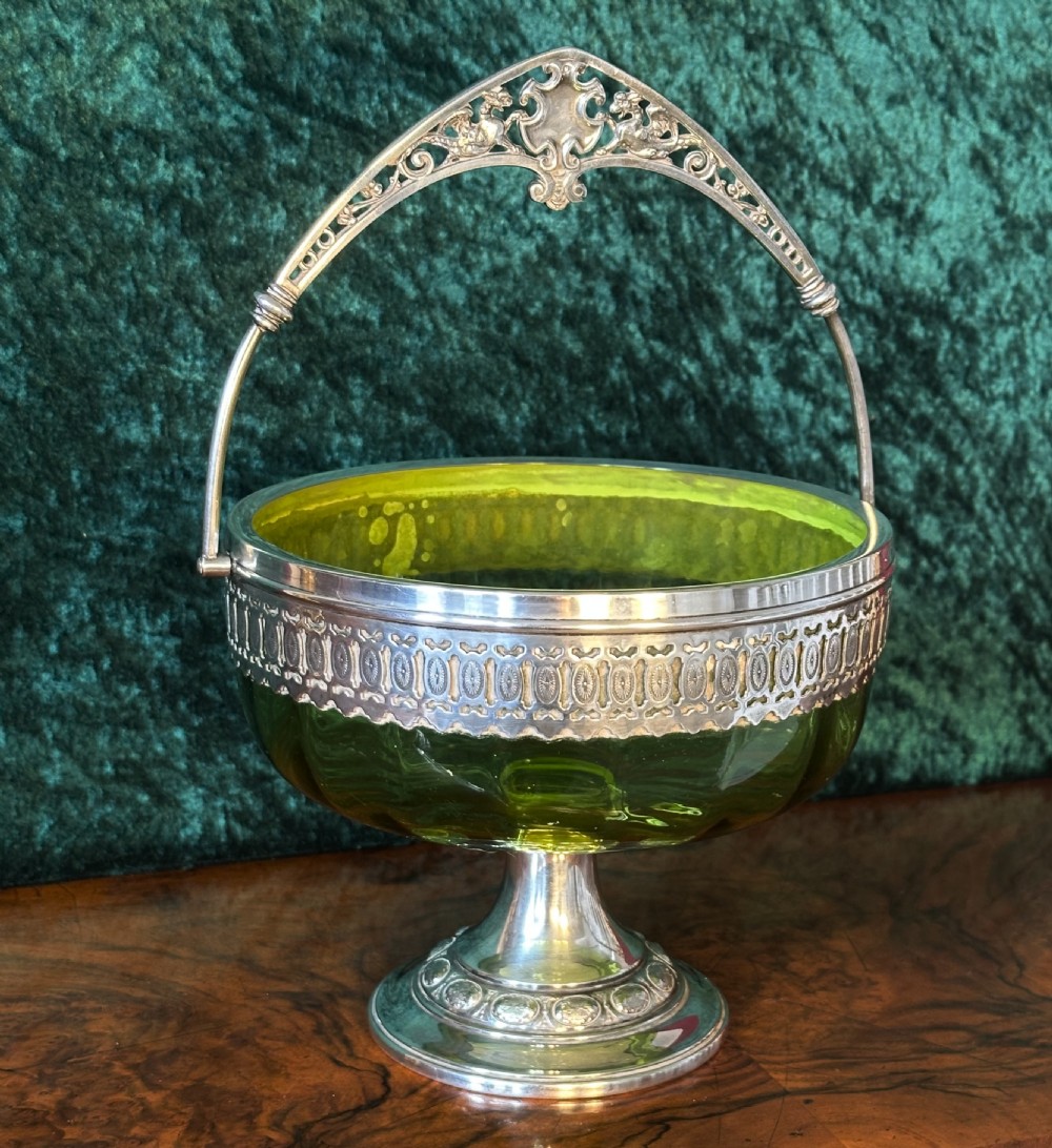 An Art Nouveau silver-plated pedestal basket featuring an ornate swing handle, pierced filigree decorative band, and a green glass liner. View IX