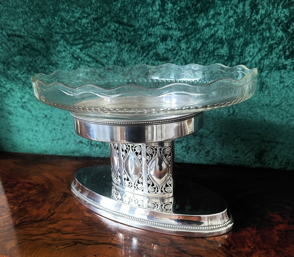 An oval silver-plated pedestal stand featuring openwork scroll panels and a clear glass dish with a scalloped rim.