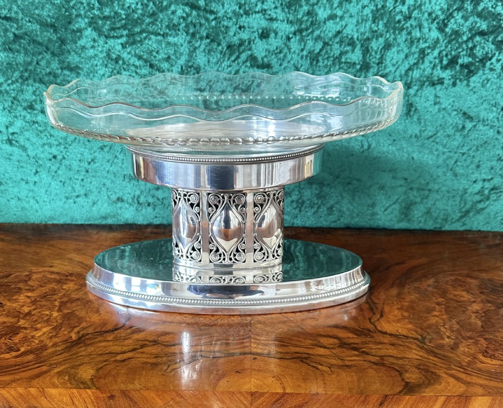 An oval silver-plated pedestal stand featuring openwork scroll panels and a clear glass dish with a scalloped rim. View II
