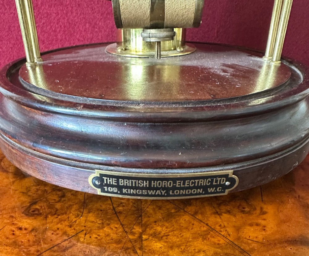 A 1920s Bulle electric clock featuring an open brass skeleton movement and white dial, encased within a clear glass dome on a dark wooden circular base. View VI