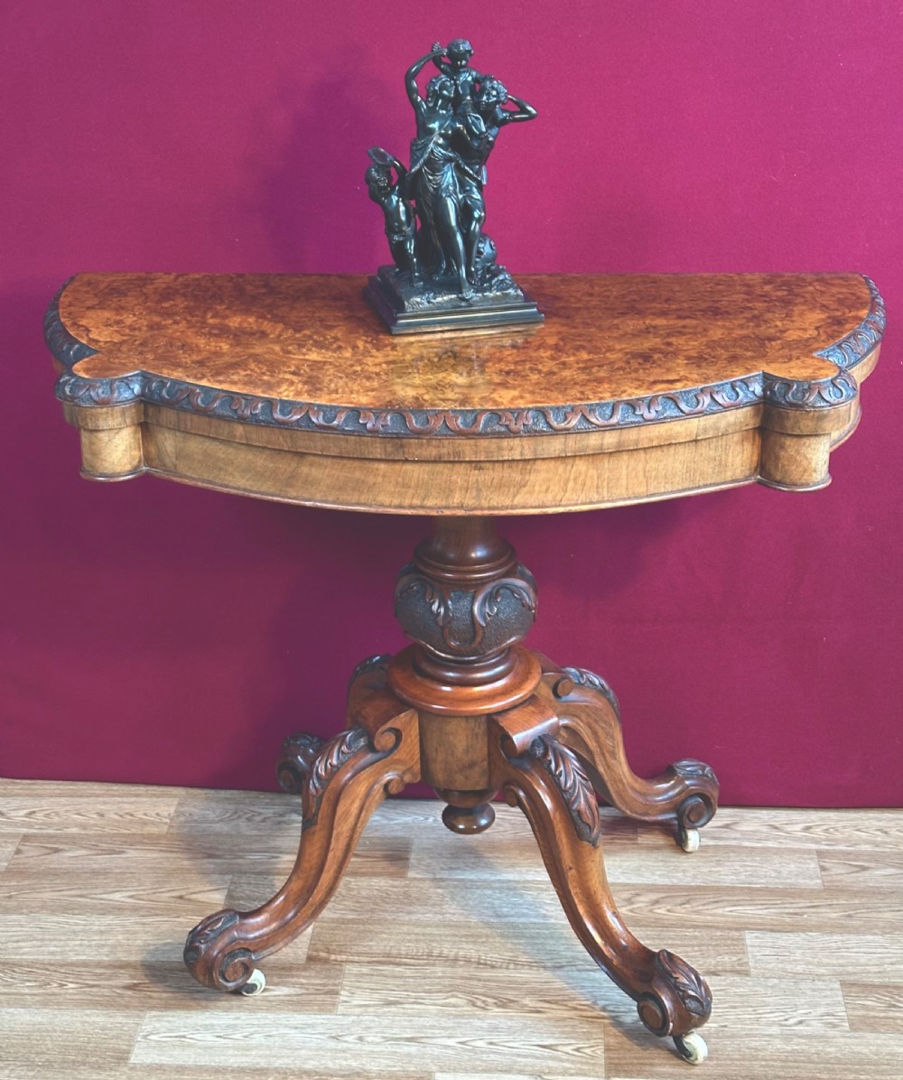 A Victorian burr walnut fold-over card table featuring a carved frieze, a central carved pedestal stem, and four cabriole legs on ceramic castors. View VII