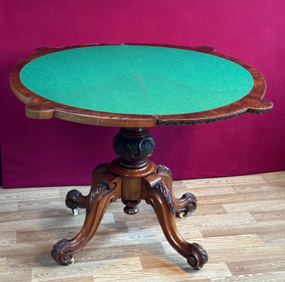 A Victorian burr walnut fold-over card table featuring a carved frieze, a central carved pedestal stem, and four cabriole legs on ceramic castors. View X