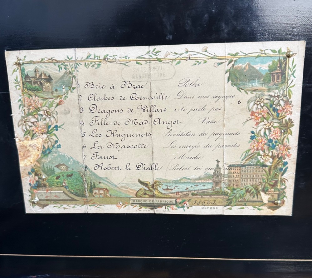 A Victorian wooden music box with an open lid displaying a printed tune sheet, brass cylinder mechanism, steel comb, and glass dust cover. View X