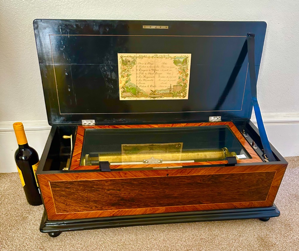 A Victorian wooden music box with an open lid displaying a printed tune sheet, brass cylinder mechanism, steel comb, and glass dust cover. View XI