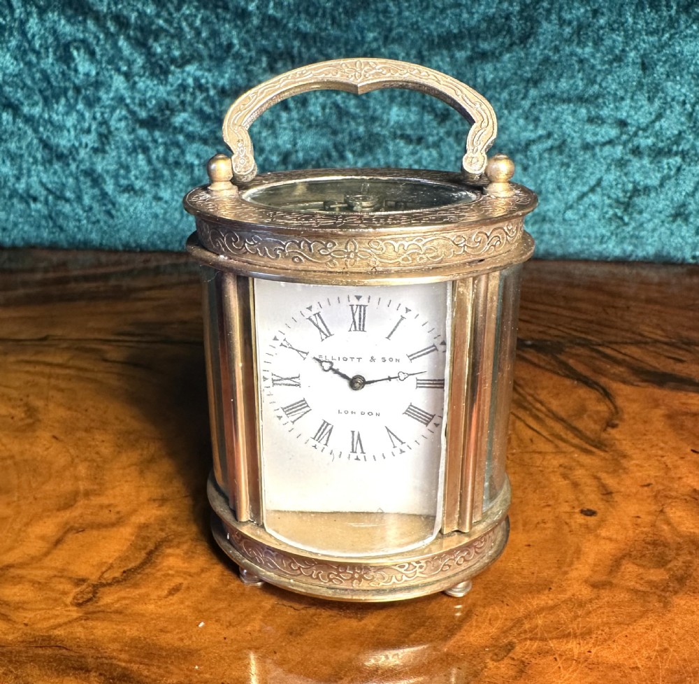 A miniature brass carriage clock featuring engraved scrollwork borders, an overhead handle, curved glass sides, and a white dial with Roman numerals signed Elliott & Son London.